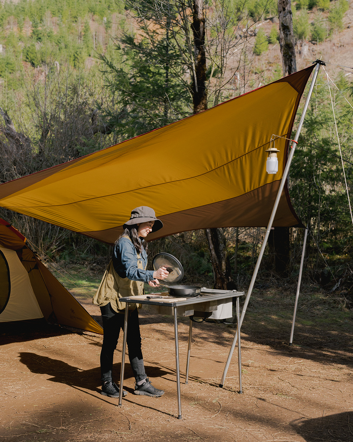 A person cooking outdoors under a yellow tarp using a camp cooking kit in a well-organized camp kitchen setup surrounded by a forest.