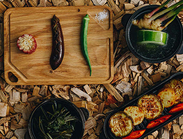 A wooden board with raw ingredients sits to the left of cast iron pots filled with cooked food. All sit on a bed of wood chips.
