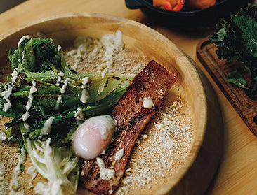A plate with bacon, poached egg, and greens sit to the left of a board filled with meats and greens.