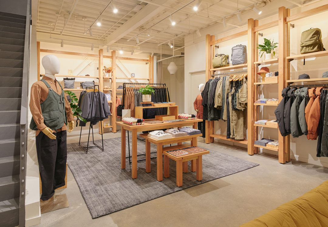 The apparel shop of the Snow Peak Brooklyn location with clothing hanging on wall racks and displayed on a table set-up in the center of the room.